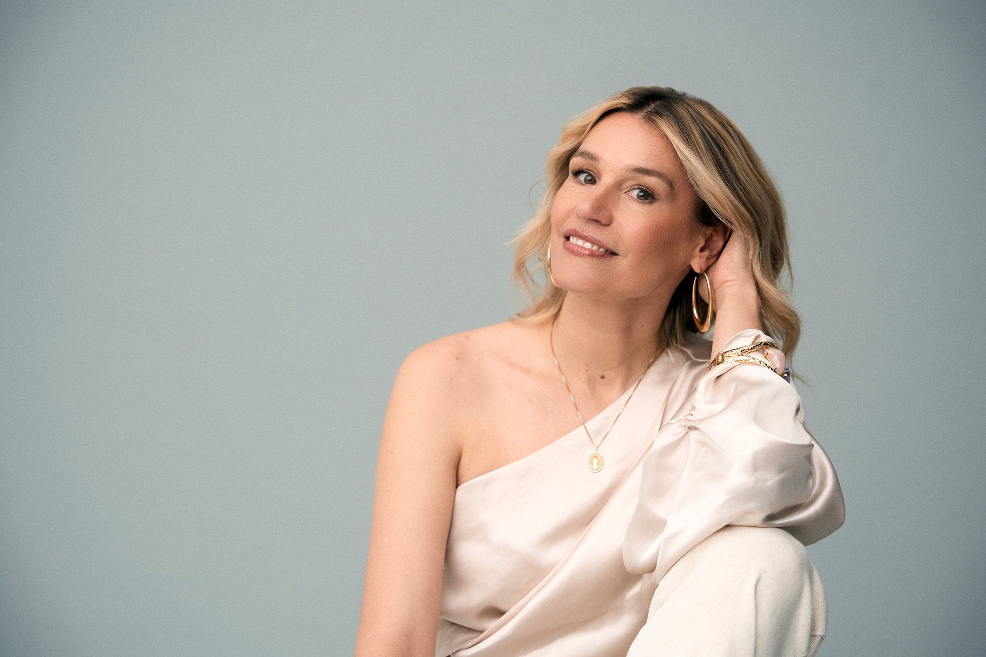 Bianca Berding, presenter, seated in a cream one-shoulder satin top against a pale blue backdrop, hand in her hair, 2025, photographed by Kilian Amrehn