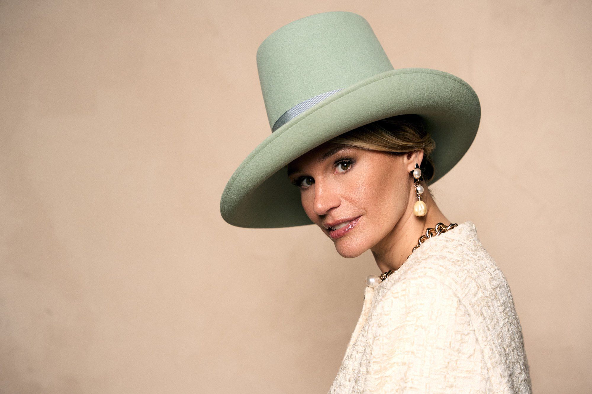 Bianca Berding, presenter, wearing a mint green hat, pearl earrings and a bouclé jacket against a beige wall, 2025, photographed by Kilian Amrehn