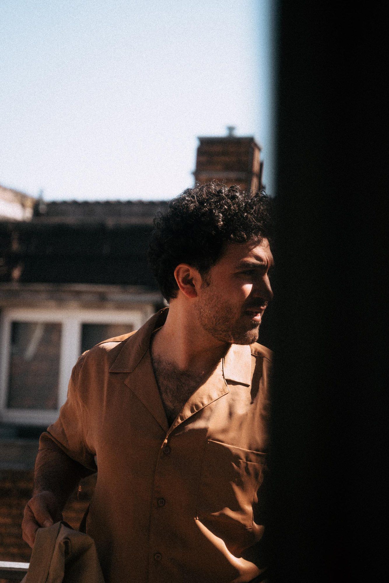 David Vormweg, actor, with curly hair in a beige shirt by a window, looking aside, hard sunlight, 2021, photographed by Kilian Amrehn