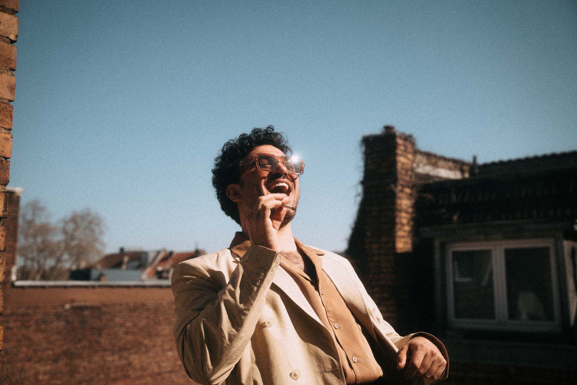 David Vormweg, actor, laughing in glasses and a light blazer on a rooftop under a blue sky, 2021, photographed by Kilian Amrehn