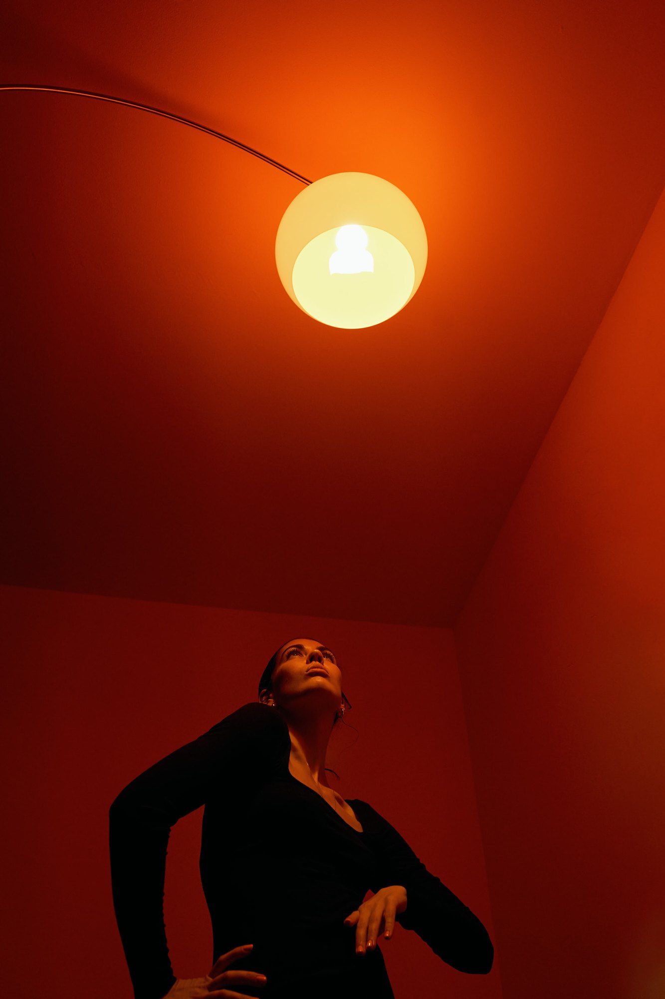 Lea Mende in black looking up at a glowing lamp, room bathed in orange-red light, 2023, photographed by Kilian Amrehn