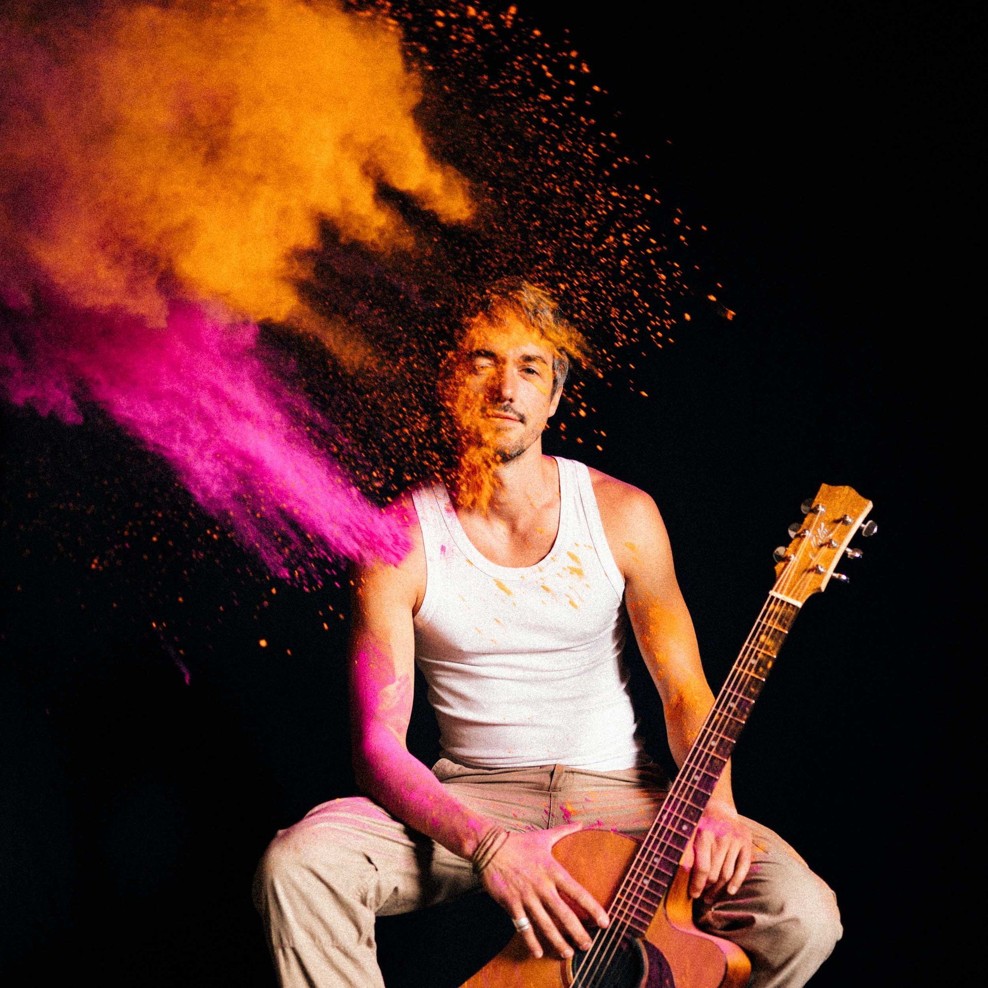 Mario Nyeky, musician, in a white tank top with an acoustic guitar, cloud of pink and orange powder against black, 2021, photographed by Kilian Amrehn