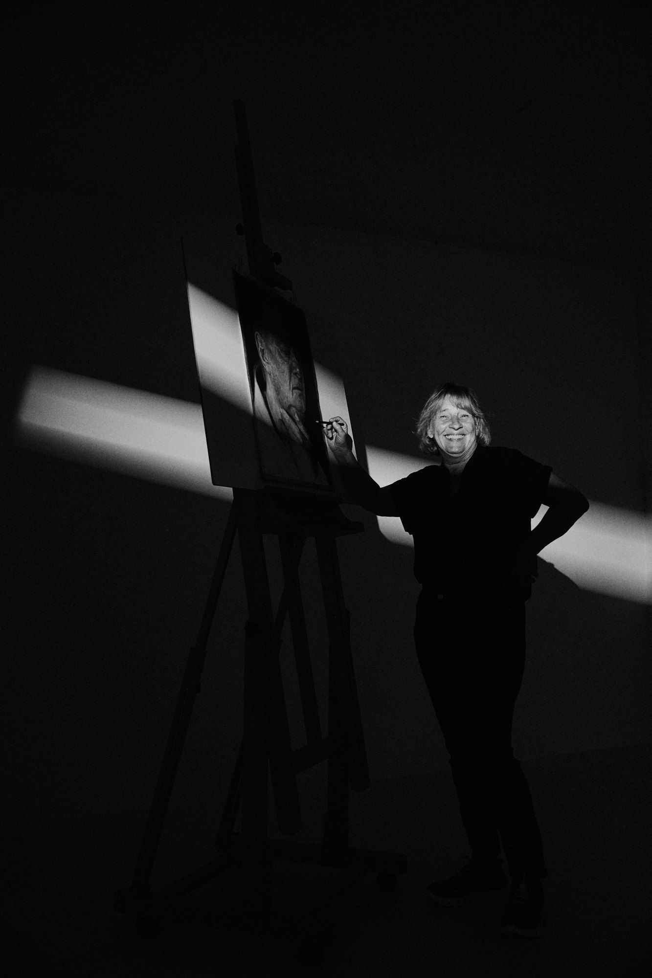 Nicole, painter, at her easel in a dark studio, a narrow strip of light across the canvas, black and white, 2026, photographed by Kilian Amrehn