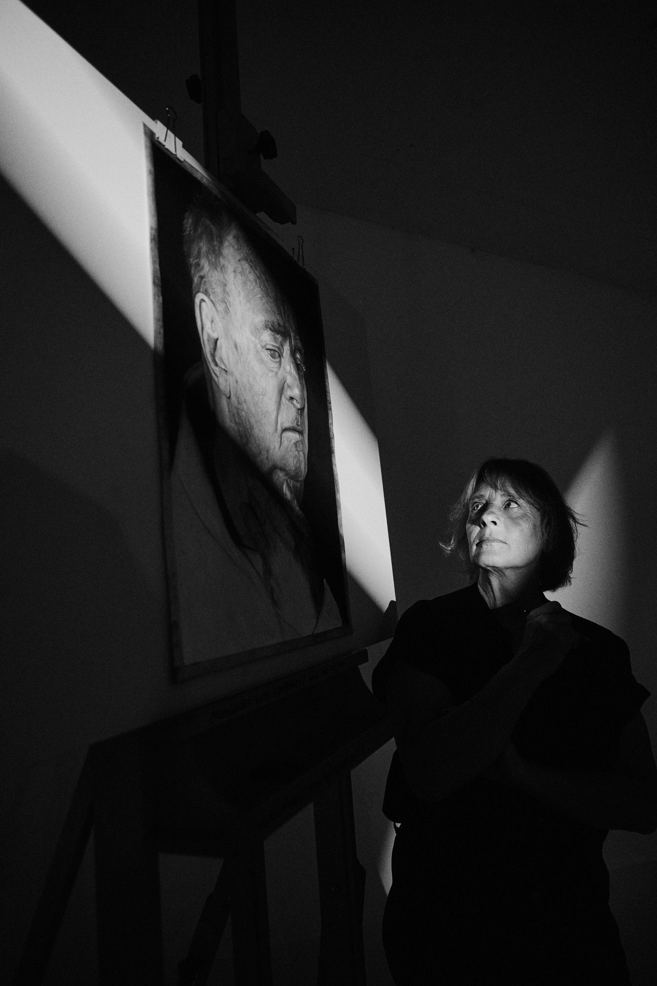 Nicole, painter, looking up at her large charcoal portrait of an old man on the easel in a shaft of light, black and white, 2026, photographed by Kilian Amrehn