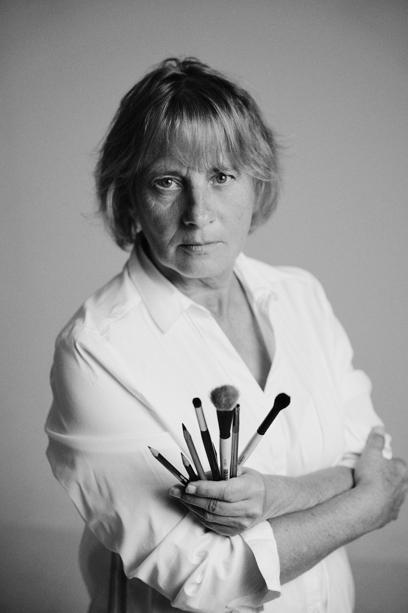 Nicole, painter, in a white shirt with folded arms holding a bundle of brushes, light background, black and white, 2026, photographed by Kilian Amrehn