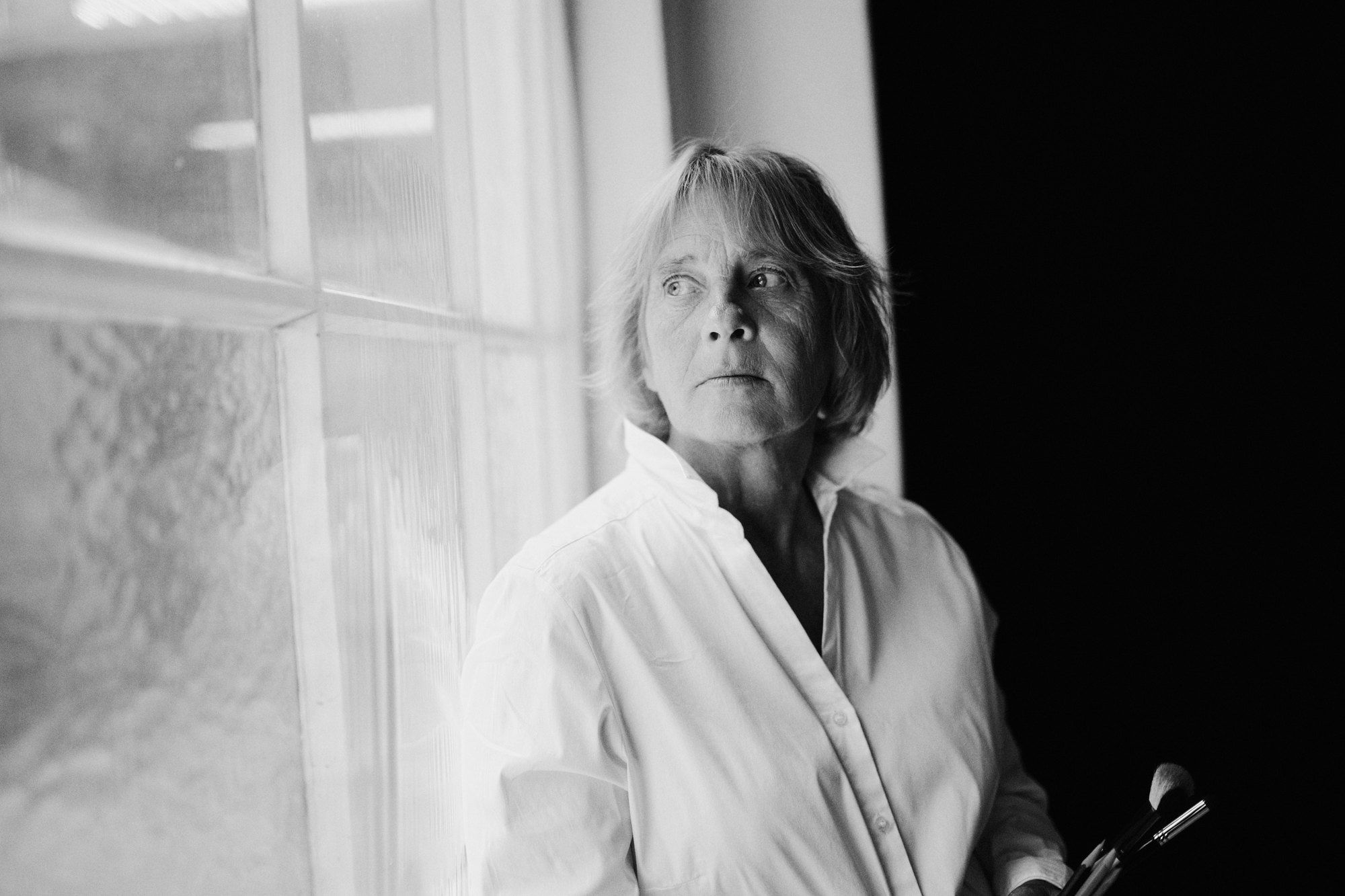 Nicole, painter, in a white shirt by a window holding brushes, looking aside, black and white, 2026, photographed by Kilian Amrehn