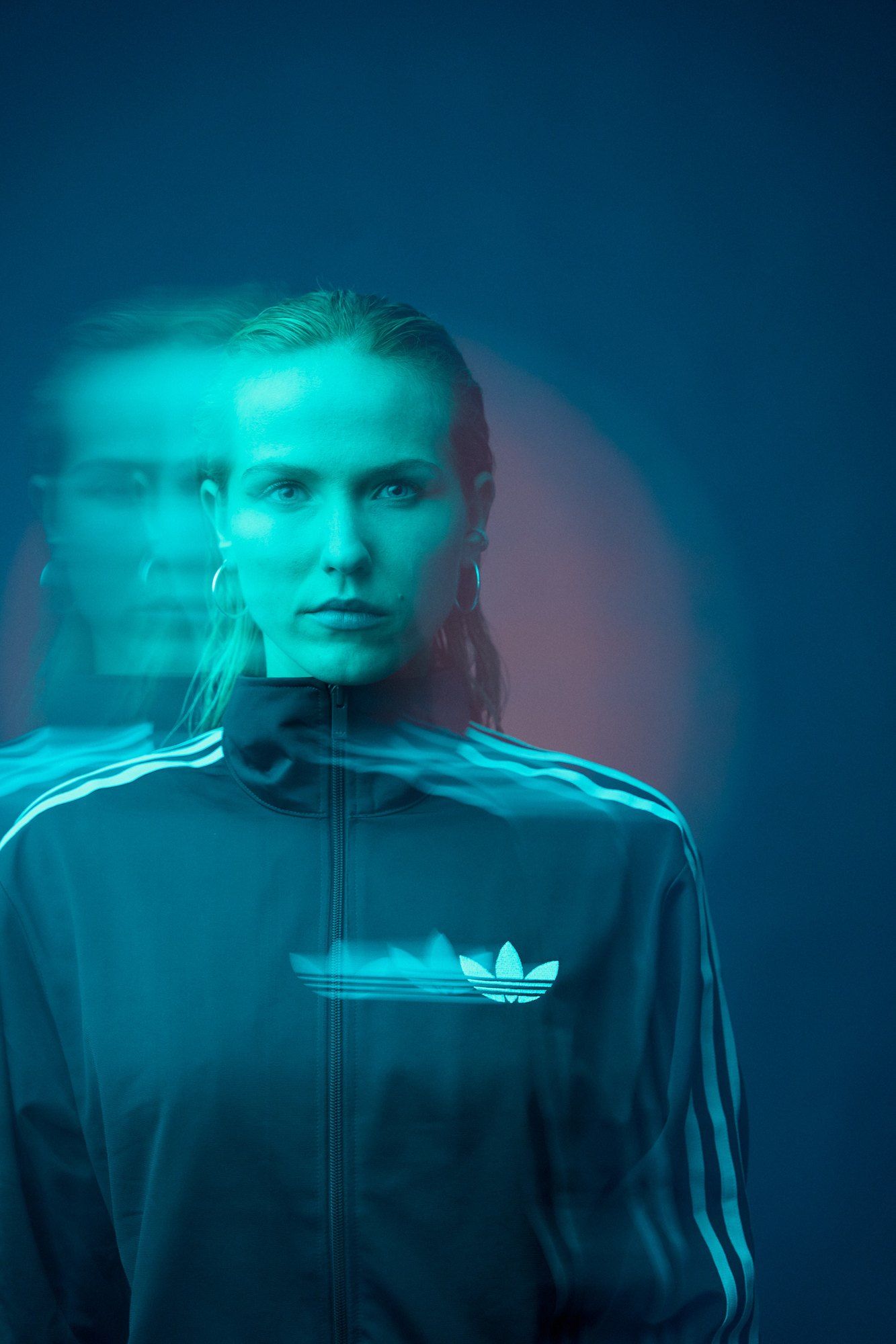 Paula, DJ, in a track jacket with an offset double image in turquoise light against a blue background, 2026, photographed by Kilian Amrehn
