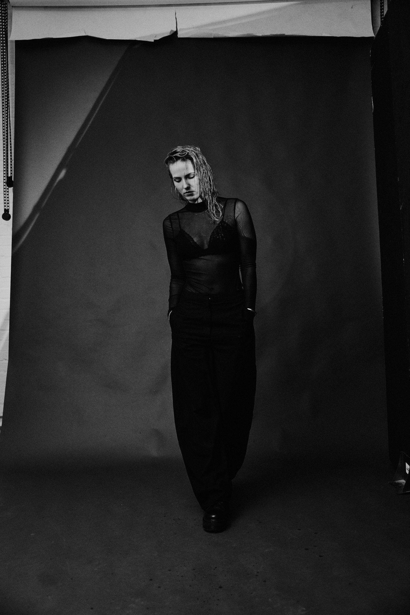 Paula, DJ, dressed all in black standing in front of a grey studio backdrop, looking down, black and white, 2026, photographed by Kilian Amrehn