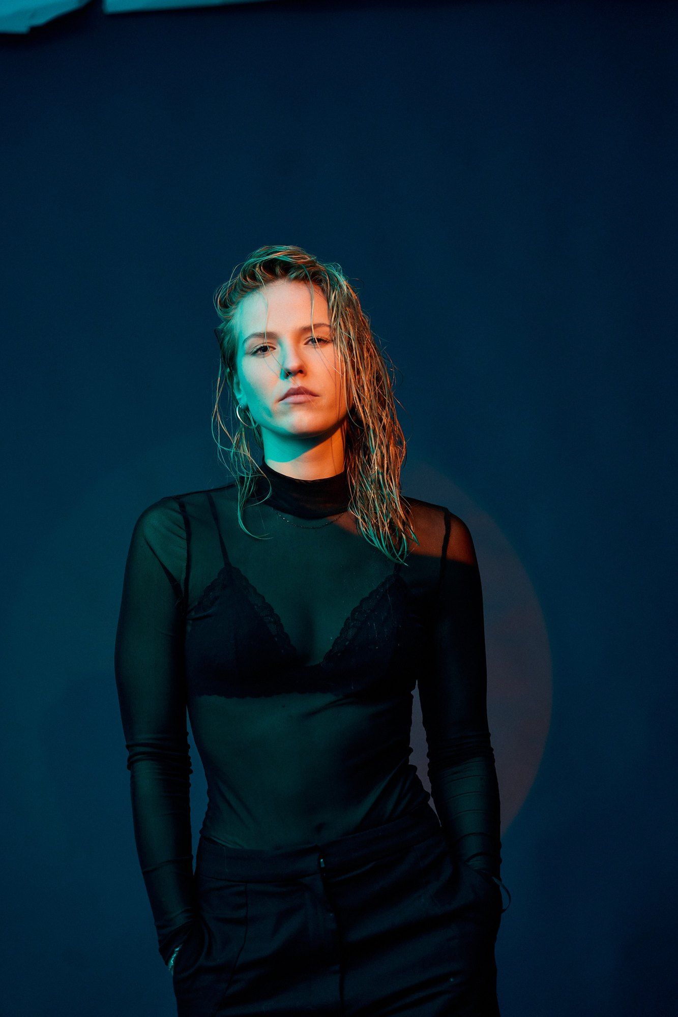 Paula, DJ, with wet hair and a sheer black top against a dark blue background, red side light, 2026, photographed by Kilian Amrehn