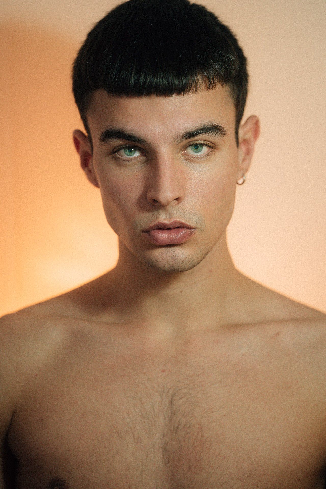 Paulus Goerden, headshot with fringe and earring, direct gaze against a warm orange background, 2022, photographed by Kilian Amrehn