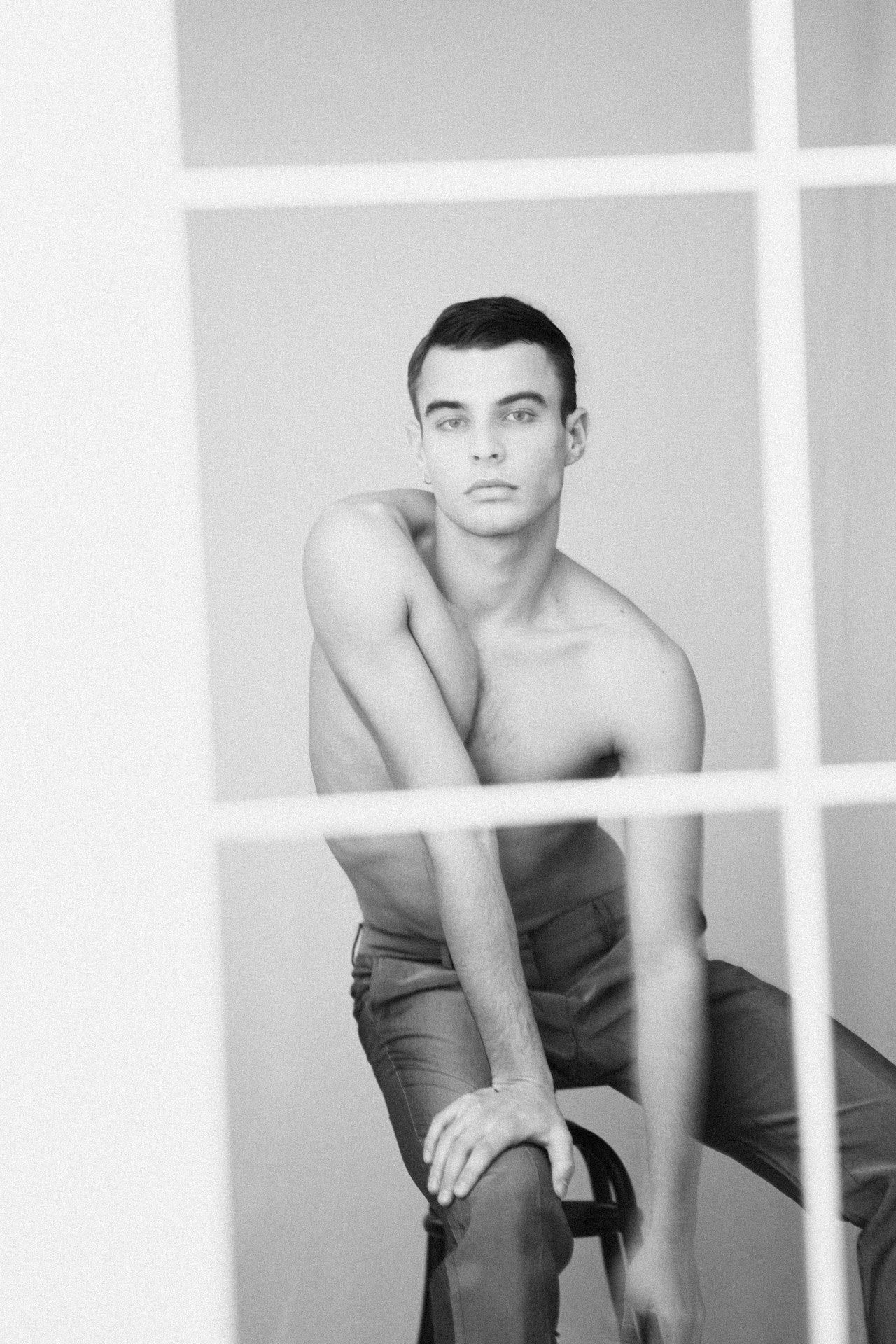 Paulus Goerden shirtless leaning on a chair behind a window frame, black and white, 2022, photographed by Kilian Amrehn