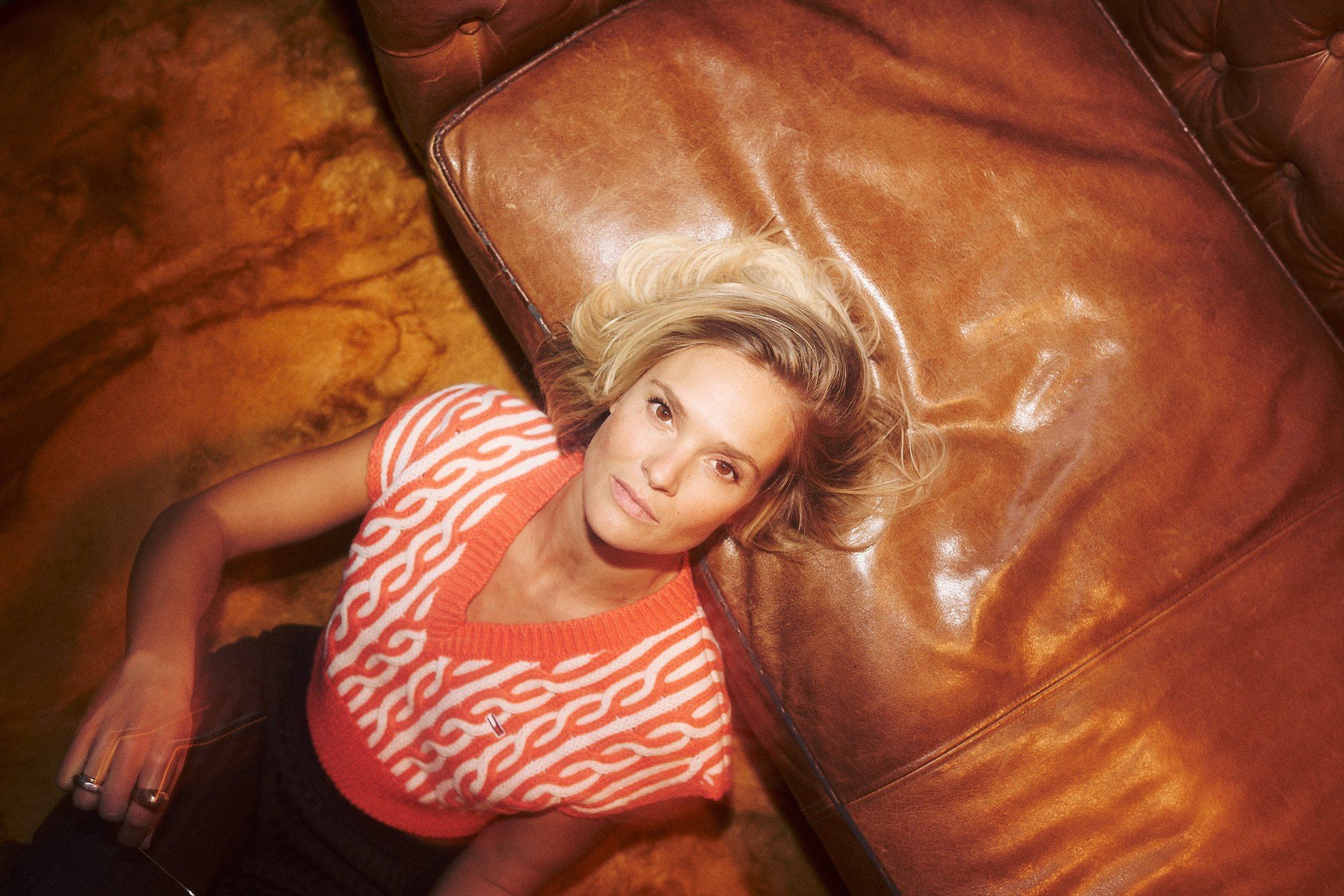 Pia Stutzenstein, actress, lying on a brown leather sofa in a red and white knitted vest, seen from above, 2022, photographed by Kilian Amrehn