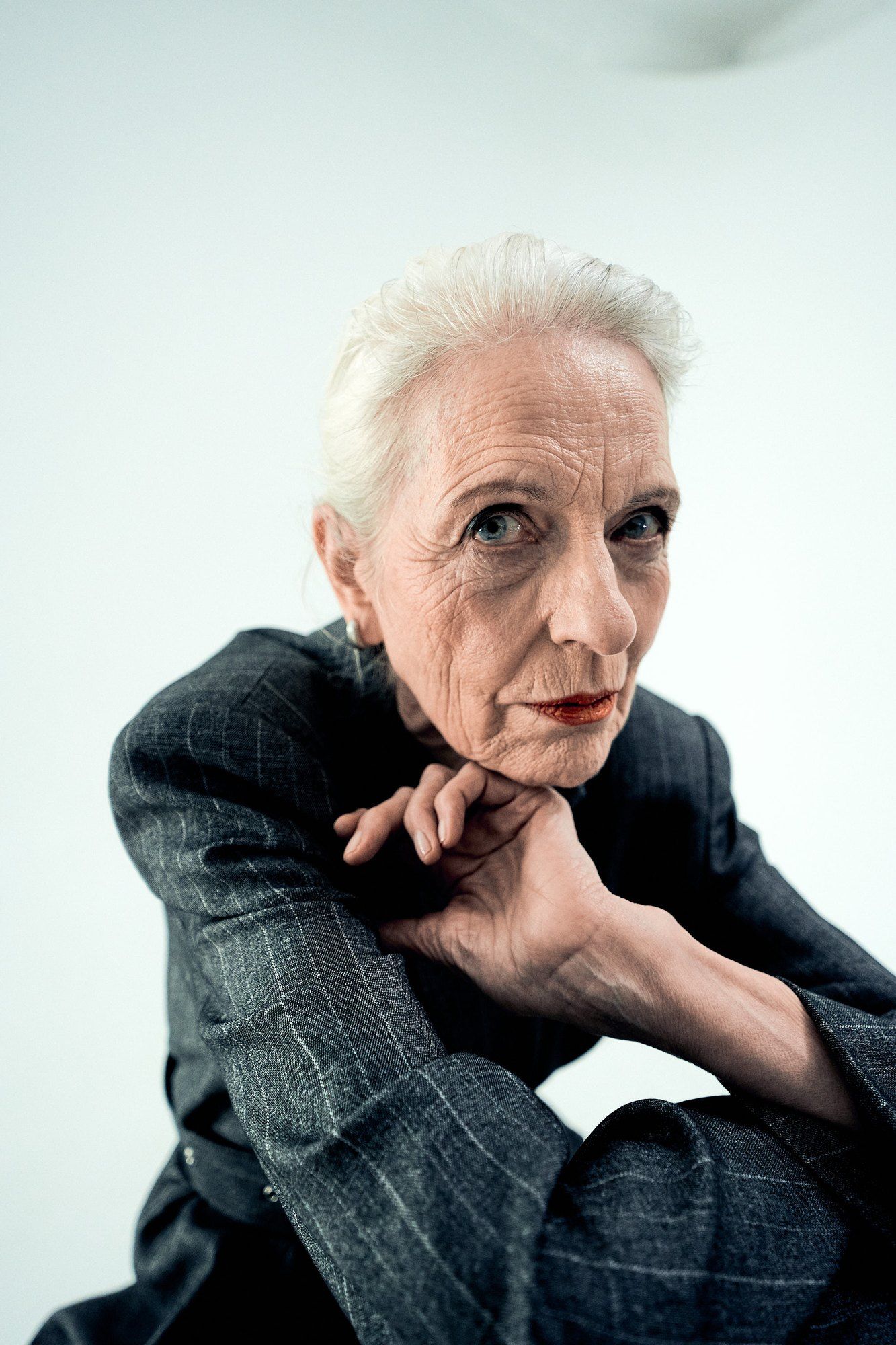 Older woman with white hair and red lipstick, chin resting on her hand, grey pinstripe suit against a light background, 2023, photographed by Kilian Amrehn