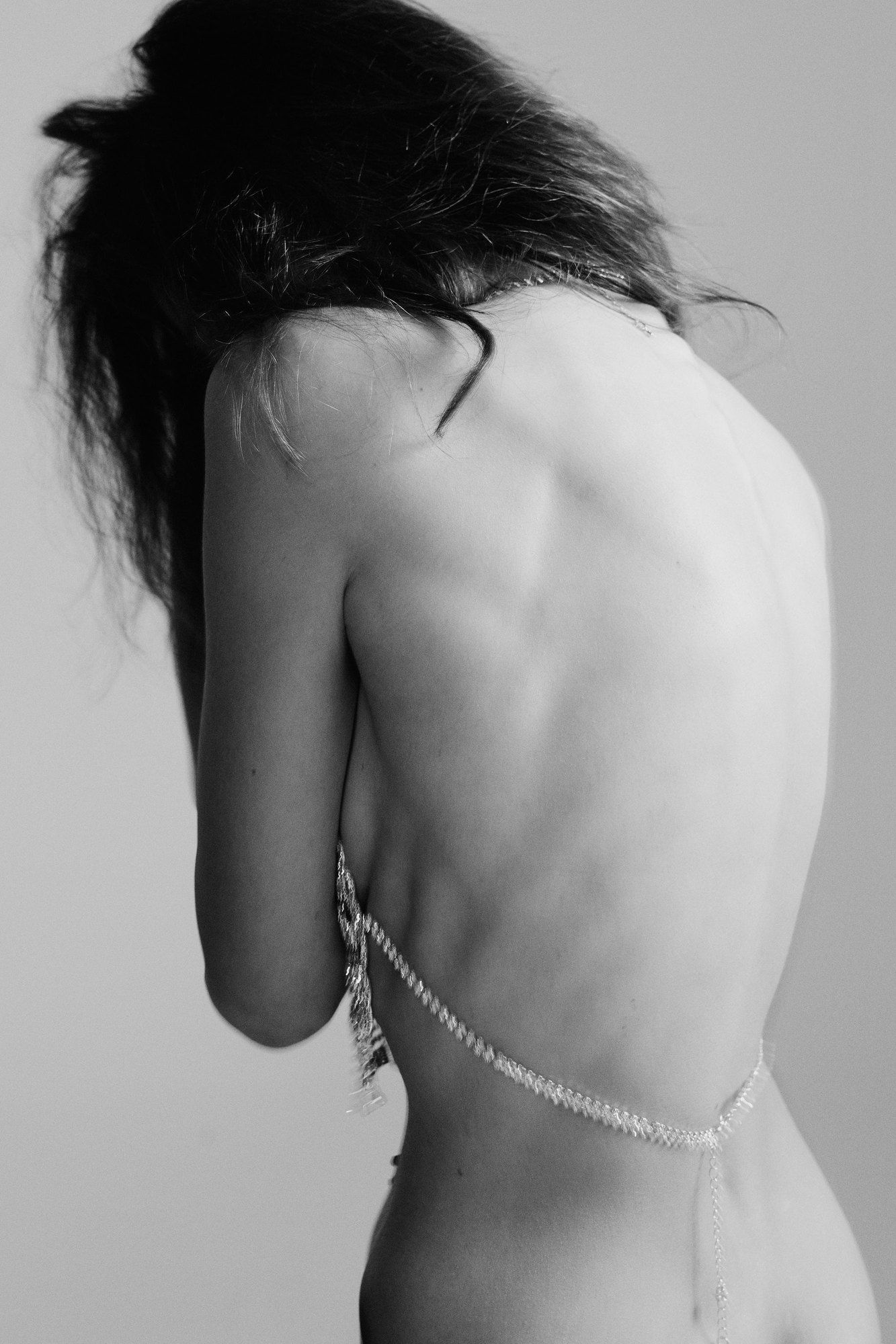 Woman with long dark hair seen from behind, backless dress with a beaded chain, light grey background, black and white, 2024, photographed by Kilian Amrehn