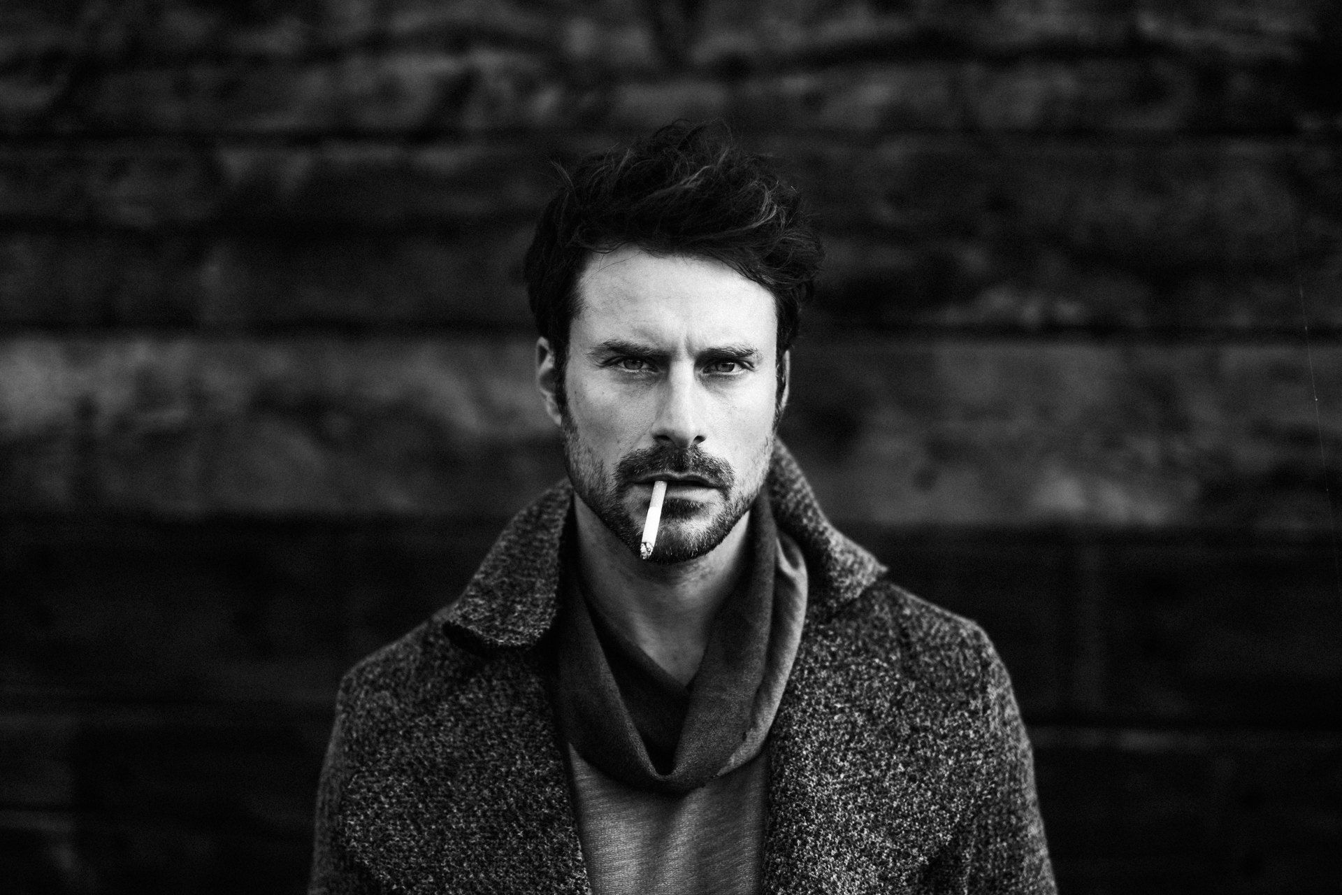 Bearded man with a cigarette in the corner of his mouth, tweed coat and scarf against a dark wooden wall, black and white, 2018, photographed by Kilian Amrehn