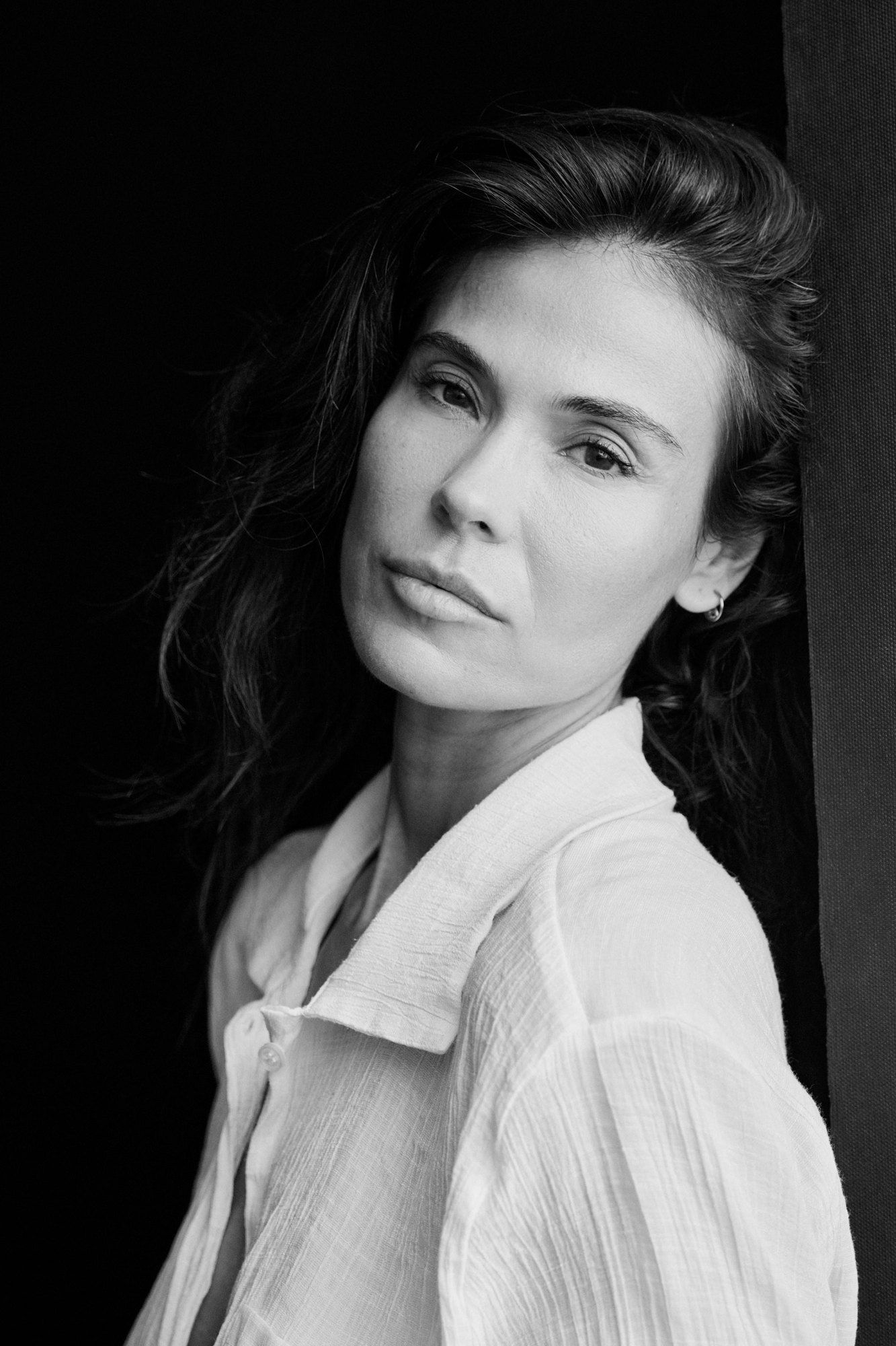 Woman with long dark hair in a white blouse leaning against a door frame, calm gaze, black and white, 2025, photographed by Kilian Amrehn