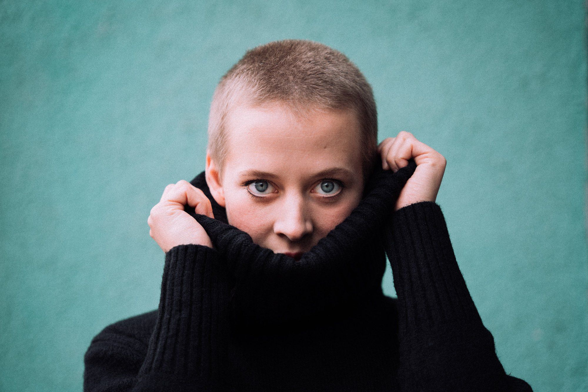 Woman with buzz-cut blonde hair pulling a black turtleneck up to her nose, in front of a turquoise wall, 2020, photographed by Kilian Amrehn