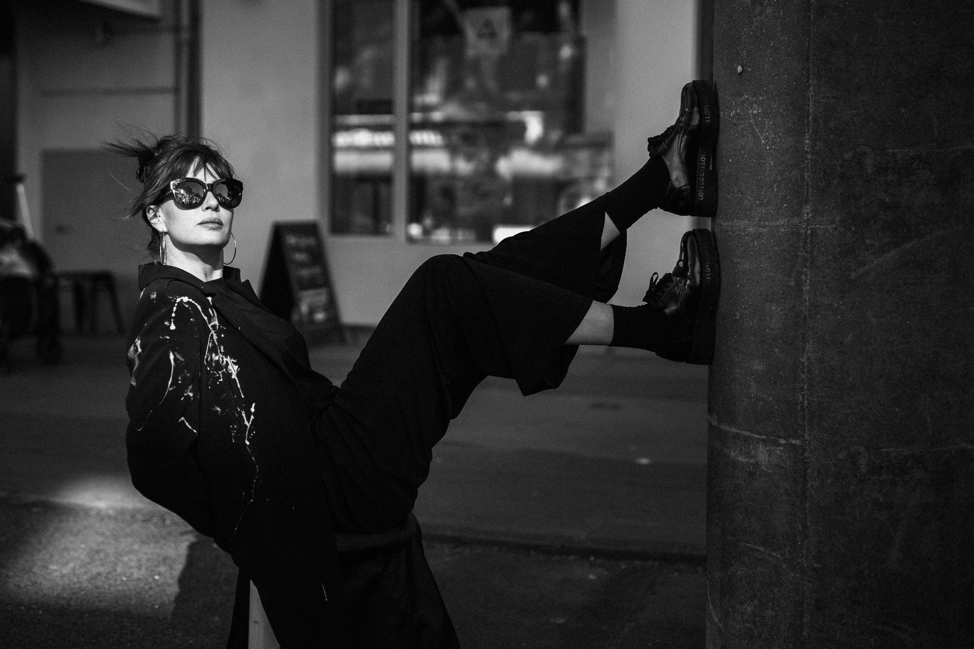 Artist in sunglasses bracing her feet against a pillar, black paint-splattered clothes on the street, black and white, 2019, photographed by Kilian Amrehn