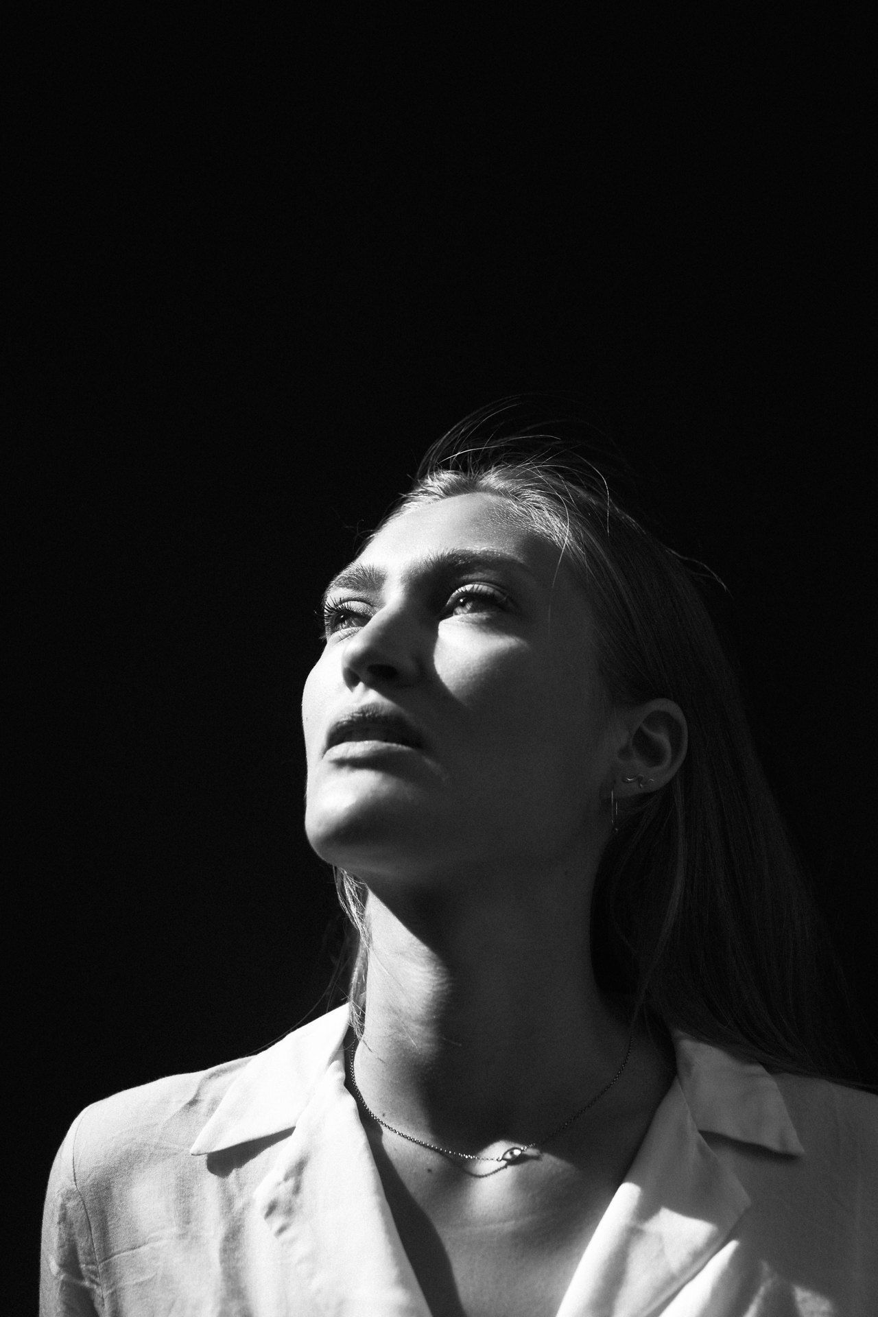 Woman with long hair in a white shirt looking up into side light against a black background, black and white, 2019, photographed by Kilian Amrehn