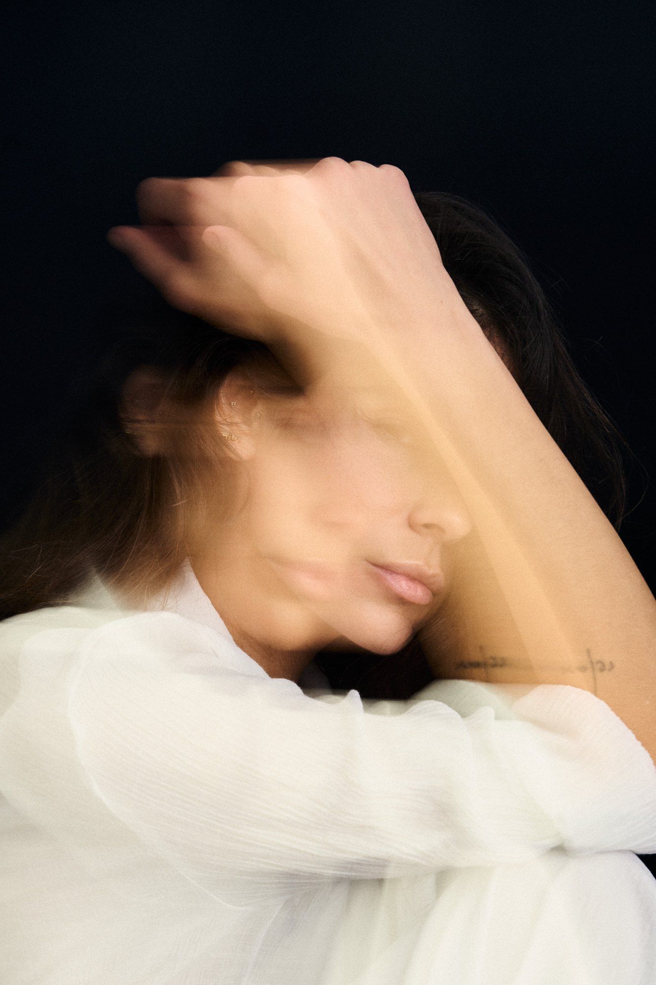 Woman in a white blouse with an arm over her head in motion blur against a black background, 2025, photographed by Kilian Amrehn