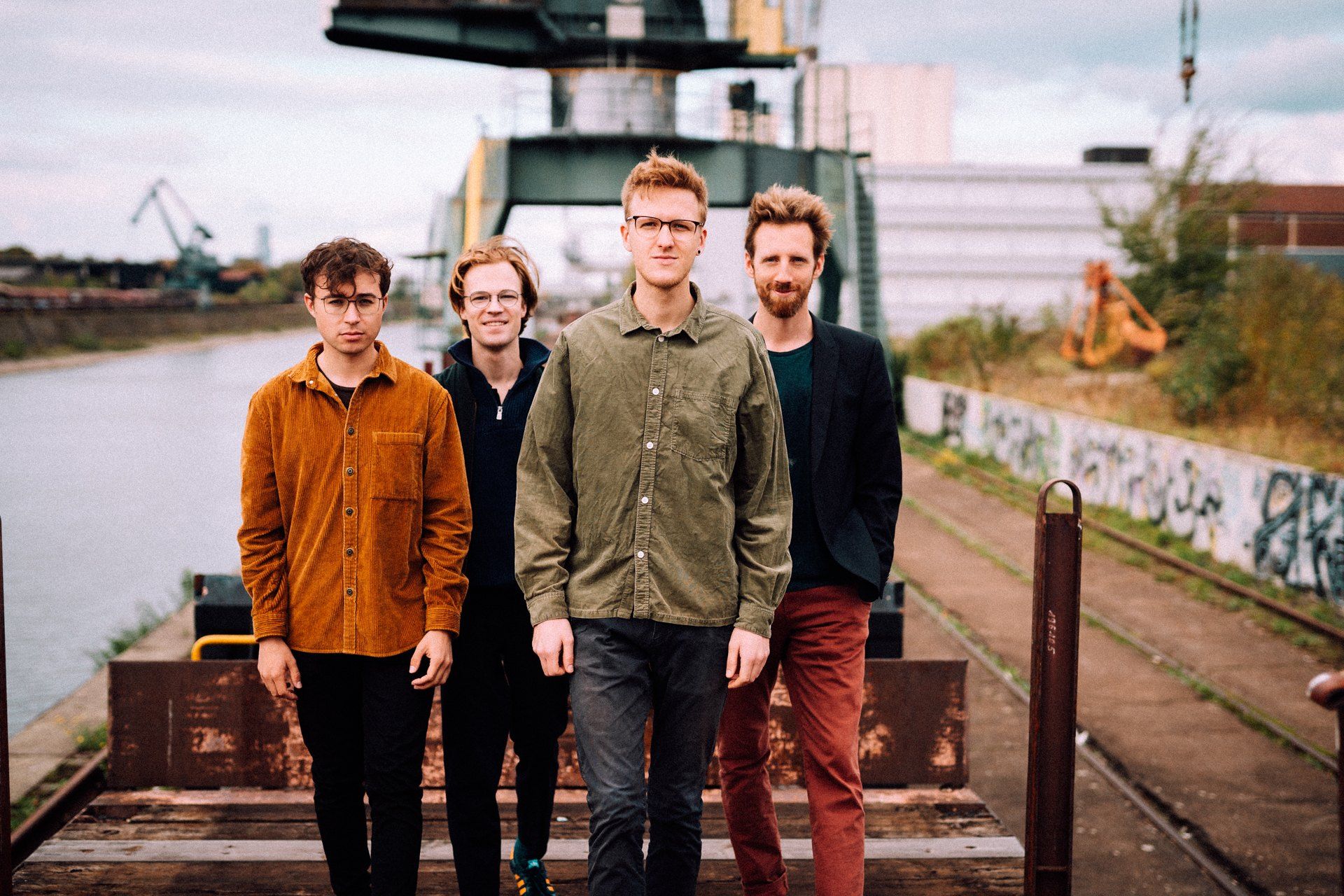 Simon und Quartett, band photo, four musicians side by side on tracks by a harbour basin, crane in the background, 2019, photographed by Kilian Amrehn