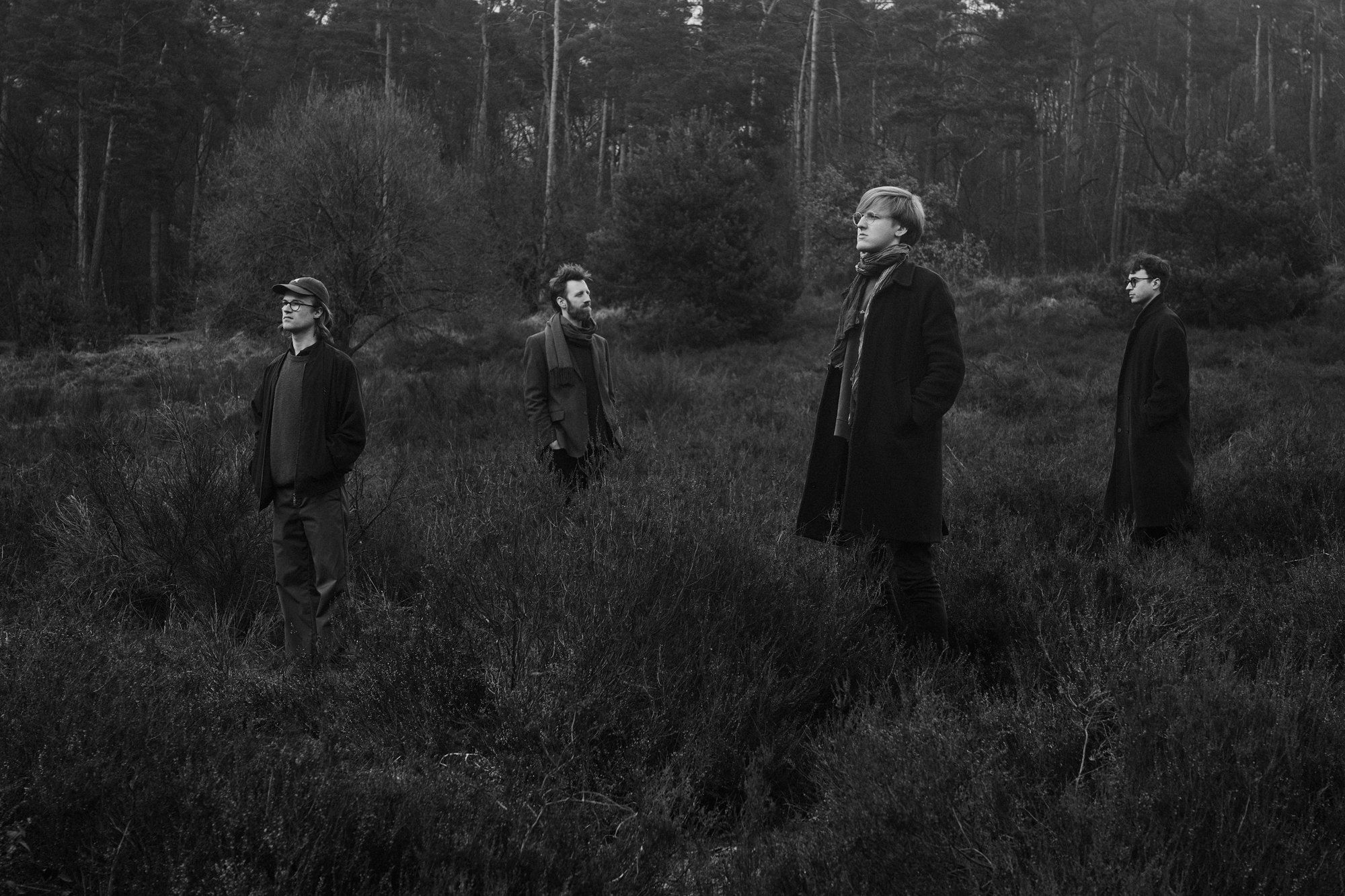 Simon und Quartett, band photo, four musicians in coats spread across heathland in front of a pine forest, black and white, 2023, photographed by Kilian Amrehn
