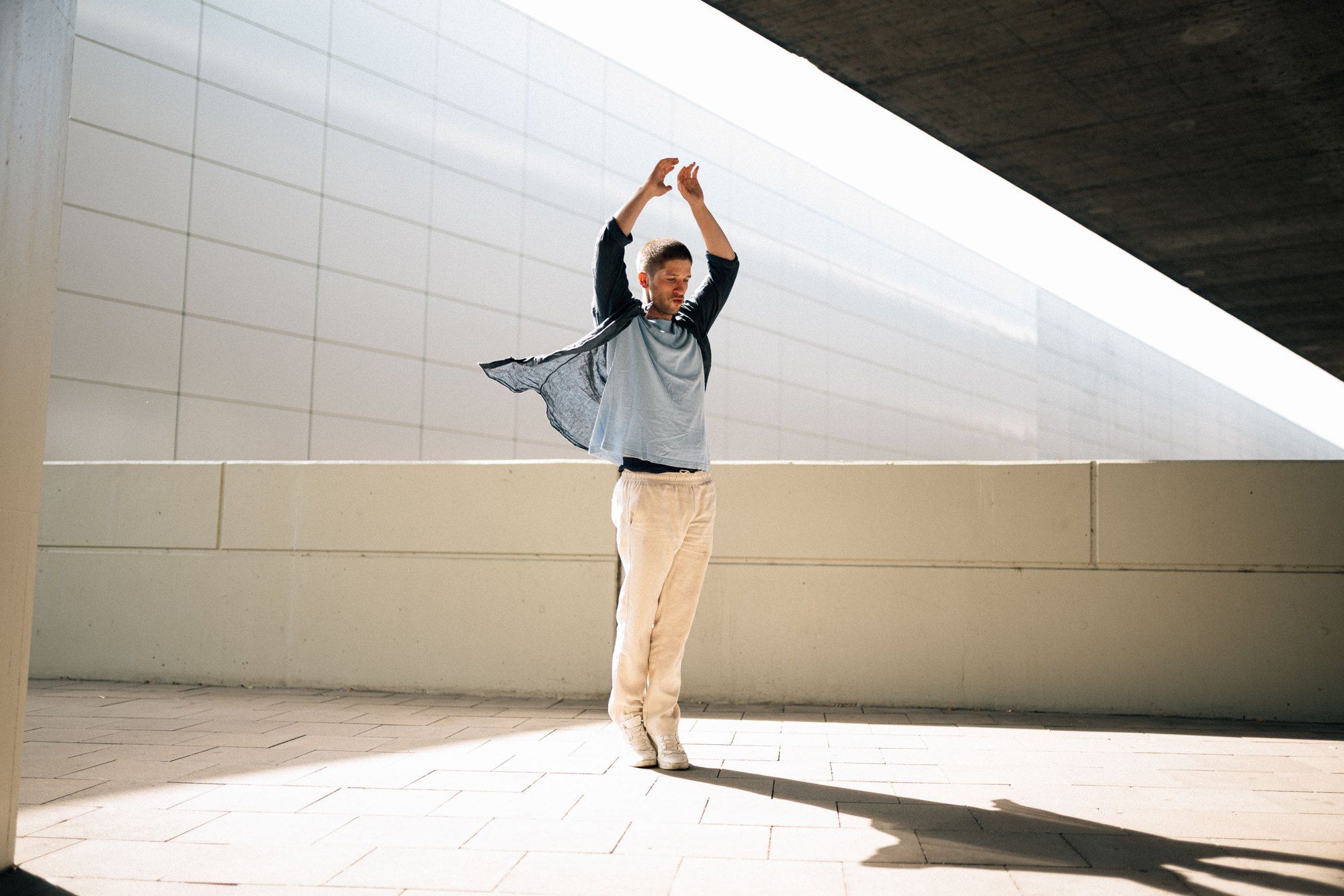 Slavko Popadić dancing with raised arms in hard sunlight beneath a concrete overhang, white trousers, long shadow, 2022, photographed by Kilian Amrehn