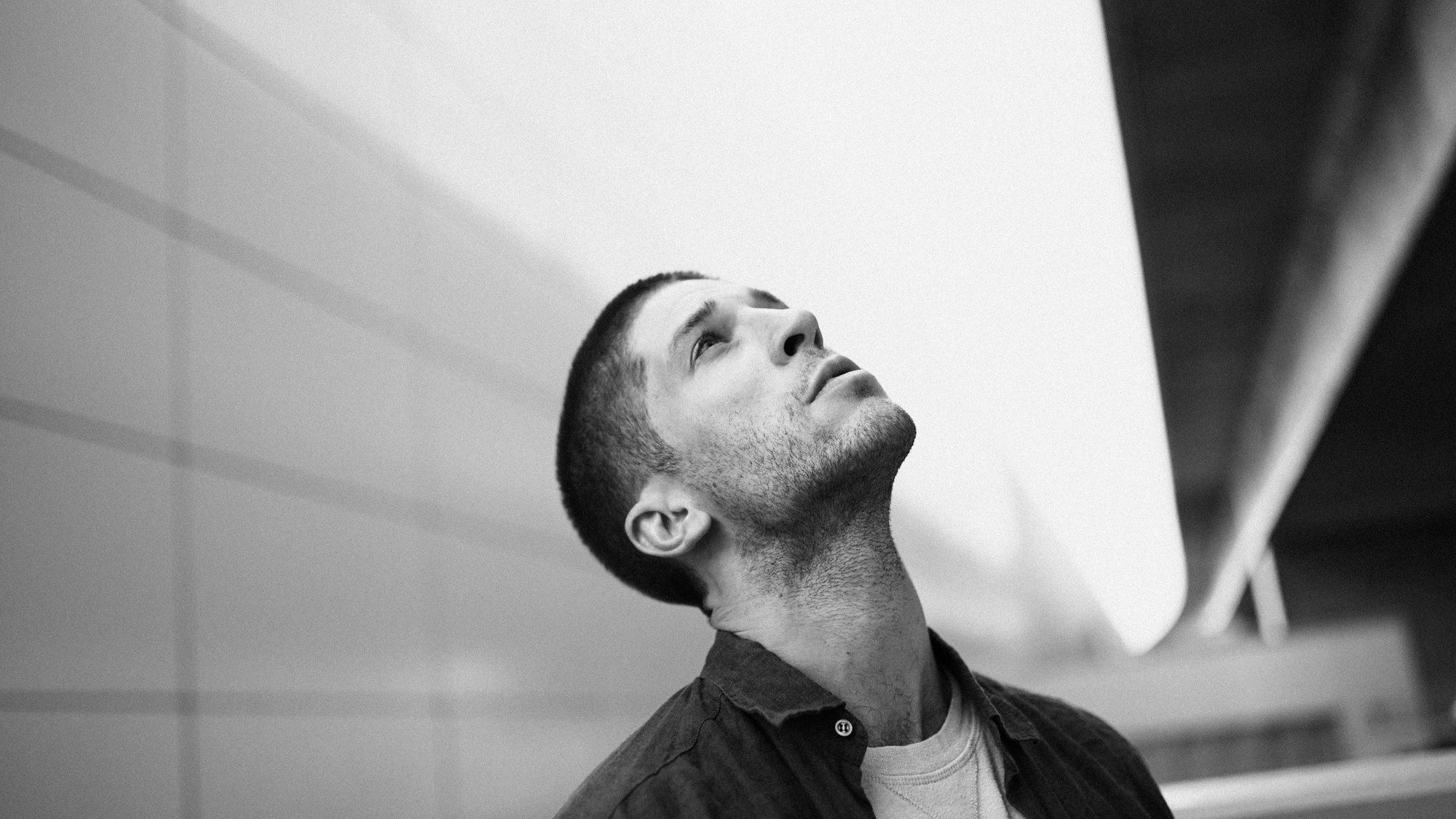 Slavko Popadić in a dark shirt looking upward in front of bright concrete architecture, black and white, 2022, photographed by Kilian Amrehn