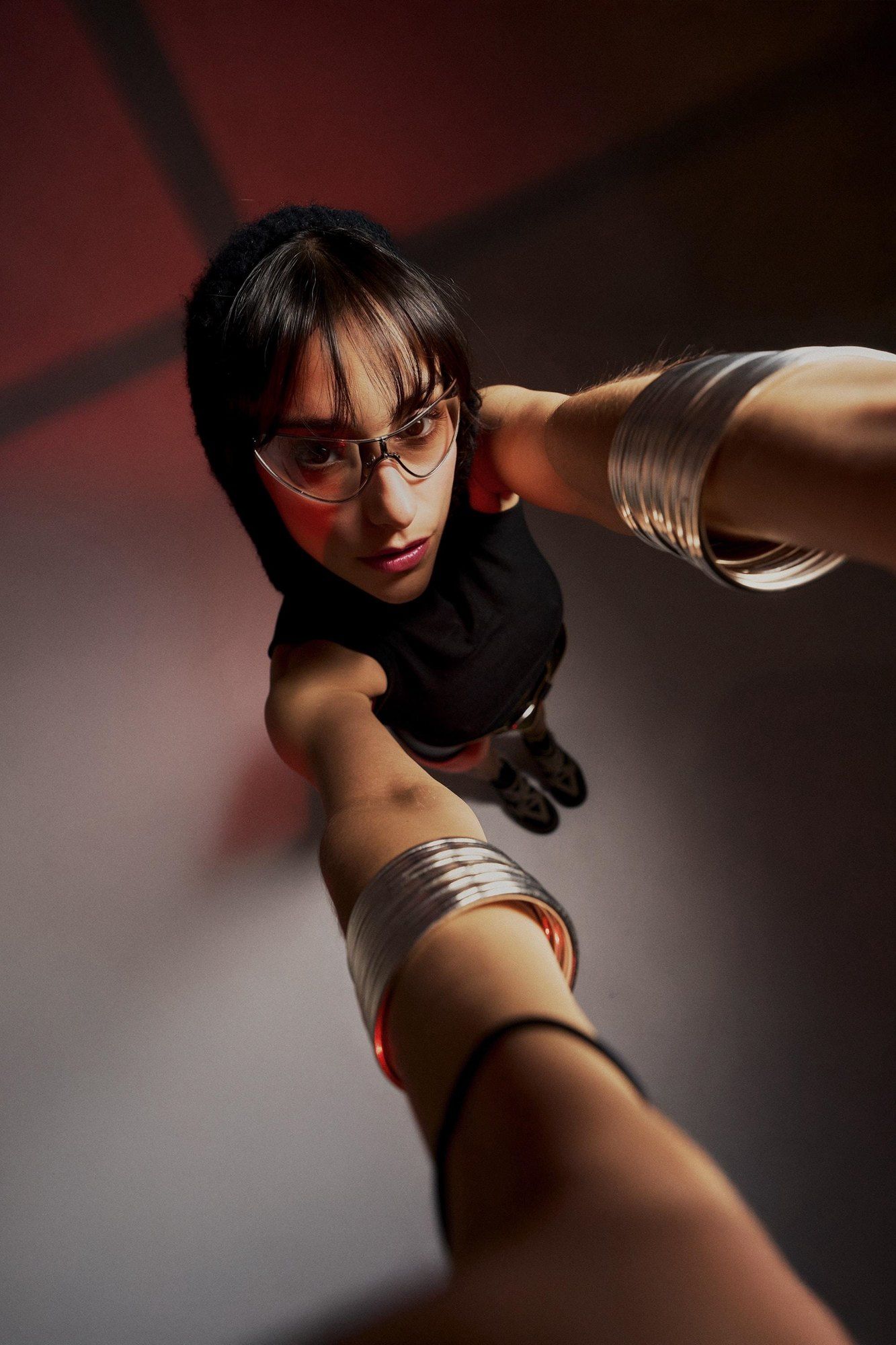 Zahnatzin, DJ, with glasses and silver bangles reaching toward the camera, wide angle from above, dark red room, 2025, photographed by Kilian Amrehn