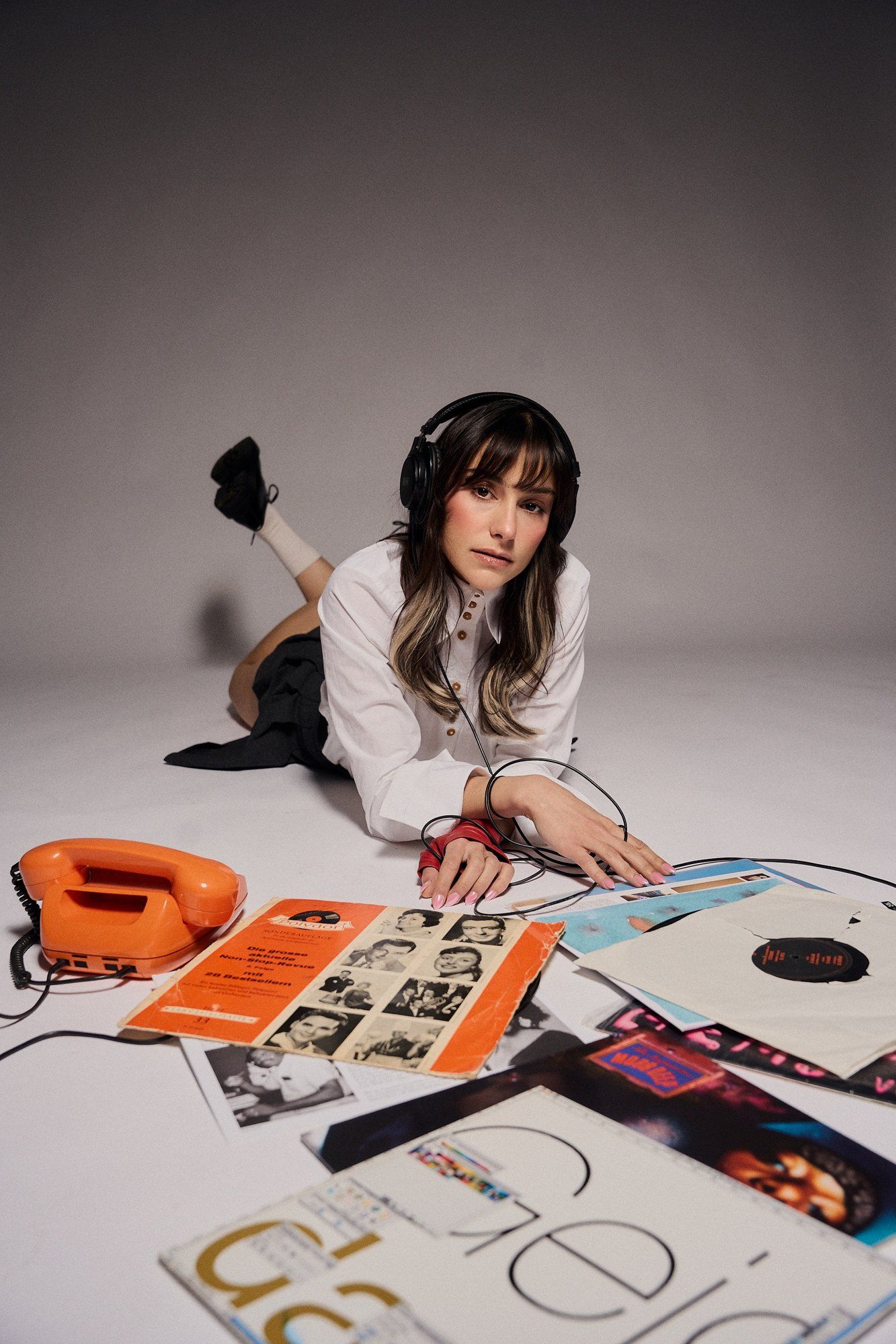 Zahnatzin, DJ, lying on the floor with headphones among vinyl records and an orange telephone, white blouse, 2025, photographed by Kilian Amrehn