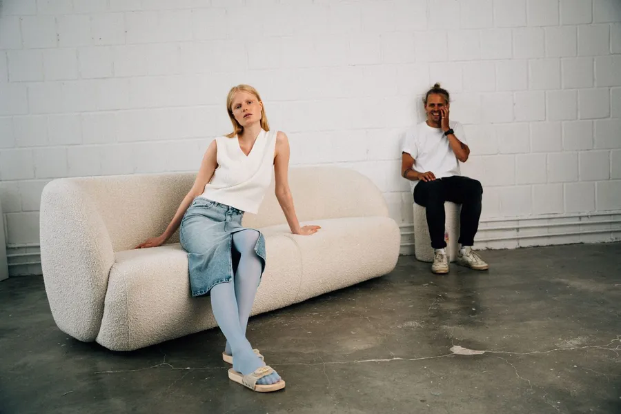Model on a white bouclé sofa in a studio, a laughing man on the phone beside her, flash by Kilian Amrehn