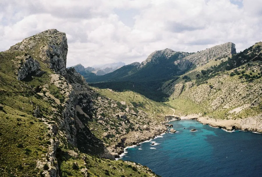 Rocky cliffs with green mountain slopes and a turquoise bay under clouds, analogue, flash by Kilian Amrehn