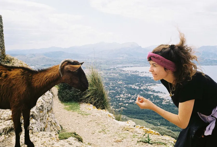 Woman in a headband leans towards a goat on a mountain path above the coast, analogue, flash by Kilian Amrehn
