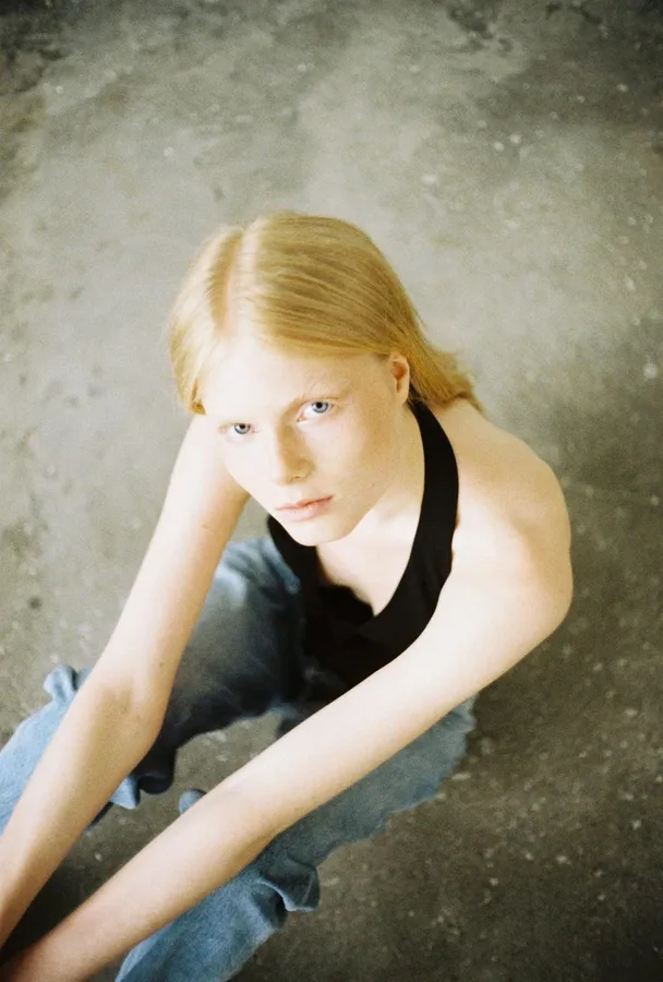Blonde woman in a halter top sits on a concrete floor looking up, analogue, flash by Kilian Amrehn