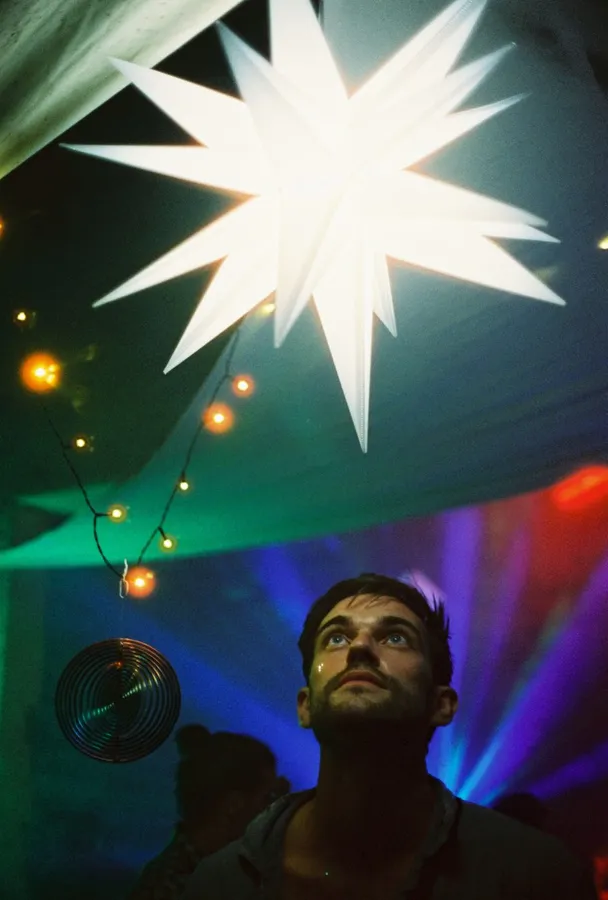 Man at a party gazes up at a glowing paper star, fairy lights and colourful lasers, flash by Kilian Amrehn