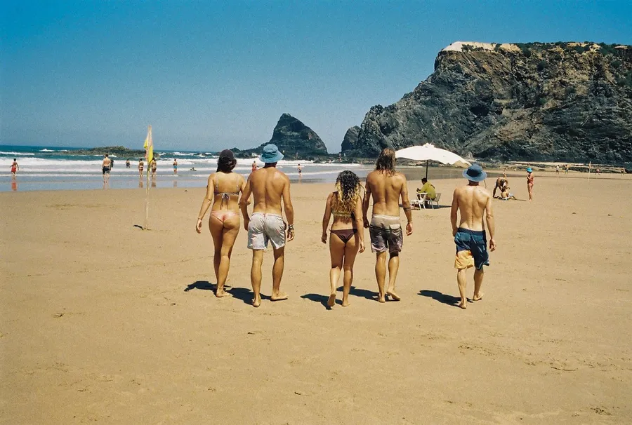 Group of friends in swimwear walks across a sandy beach towards rocks by the sea, analogue, flash by Kilian Amrehn