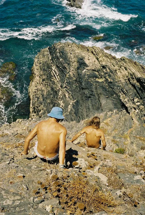 Two men sit on cliff rocks above foaming surf, analogue, flash by Kilian Amrehn