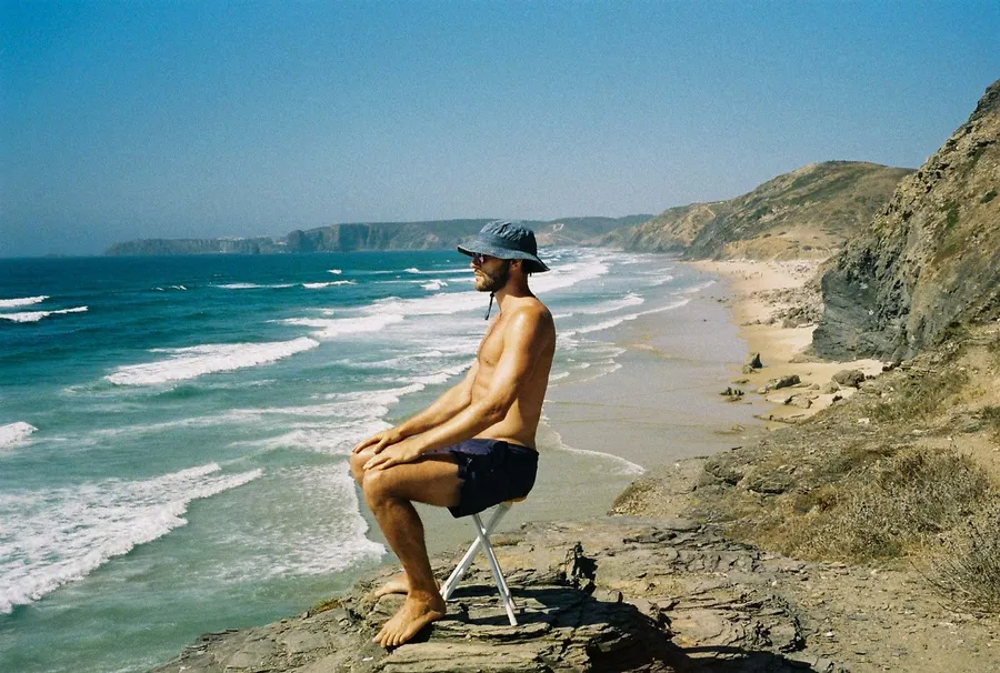 Man in a bucket hat on a stool at the cliff edge above a long surf beach, analogue, flash by Kilian Amrehn