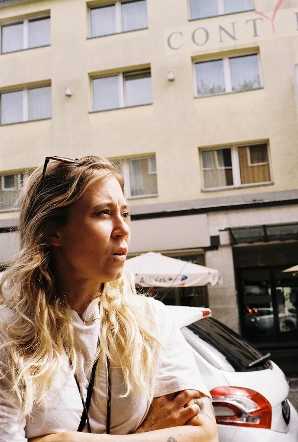 Blonde woman with sunglasses in her hair looks sideways in front of a pale facade, flash by Kilian Amrehn
