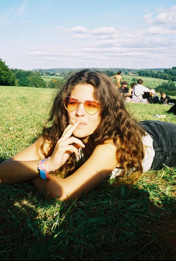 Woman in heart-shaped sunglasses lies smoking in the grass of a meadow, analogue, flash by Kilian Amrehn