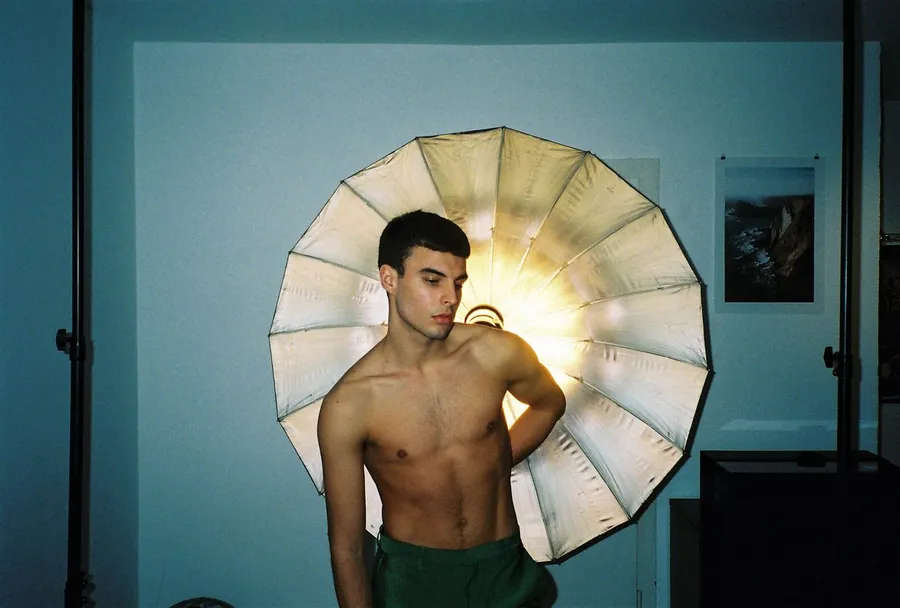 Shirtless young man in front of a silver umbrella reflector in a blue room, analogue, flash by Kilian Amrehn