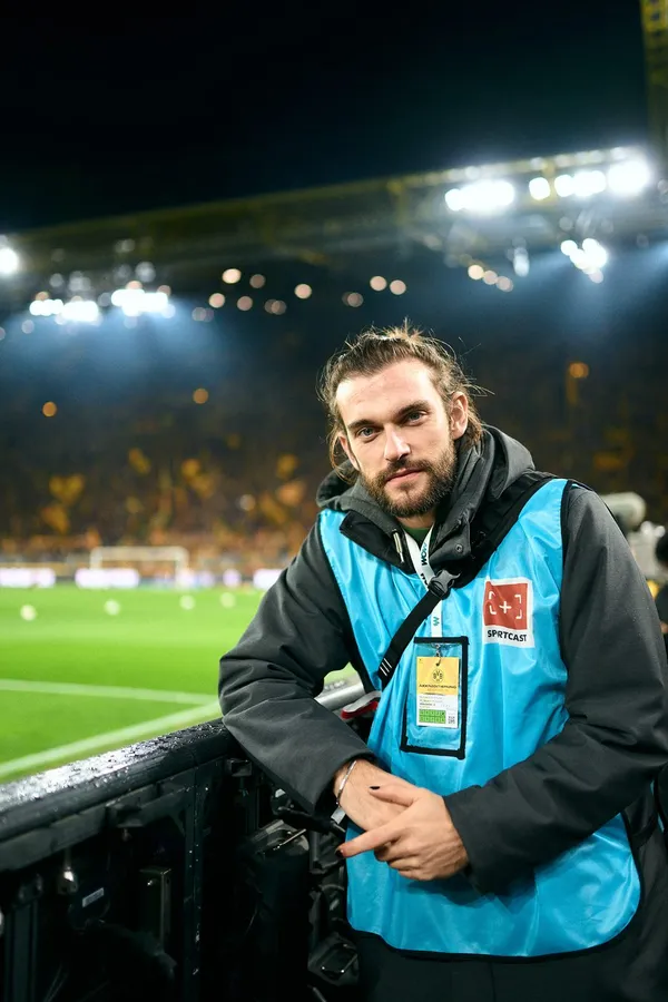 Photographer in a blue bib at the pitch side under floodlights of a packed stadium, flash by Kilian Amrehn