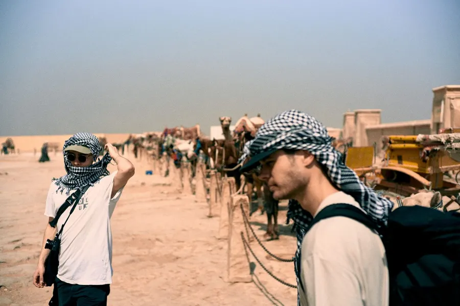 Two men in keffiyehs beside a long line of camels in the desert, flash by Kilian Amrehn