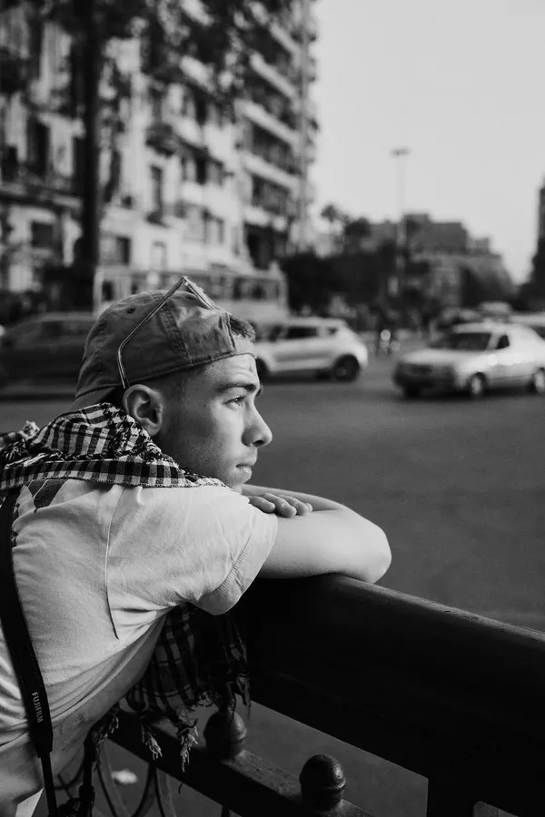 Man in a cap leans on a railing above a busy street, black and white, flash by Kilian Amrehn