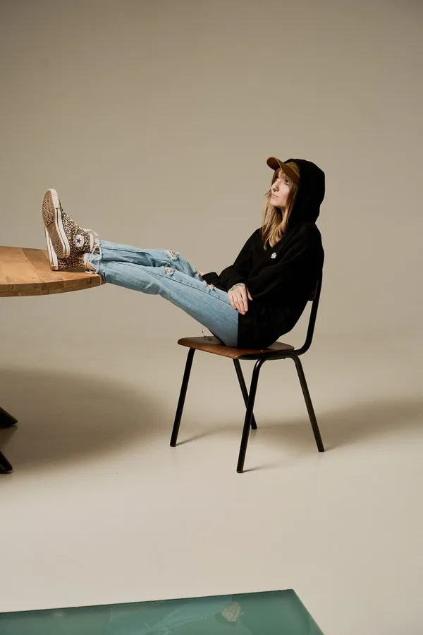 Woman in a cap rests her feet on a wooden table against a beige studio backdrop, flash by Kilian Amrehn