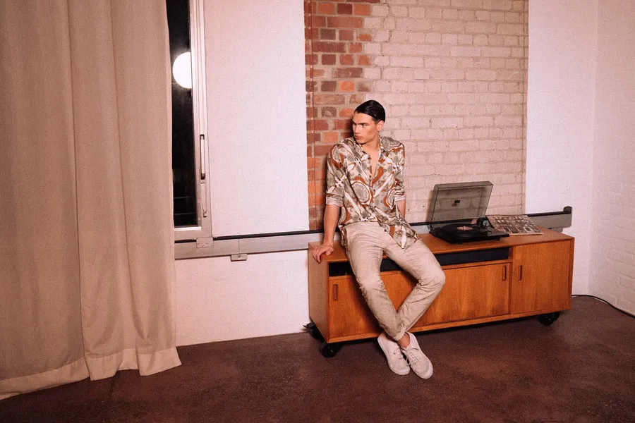 Man in a patterned shirt sits on a sideboard with a record player by a brick wall, flash by Kilian Amrehn