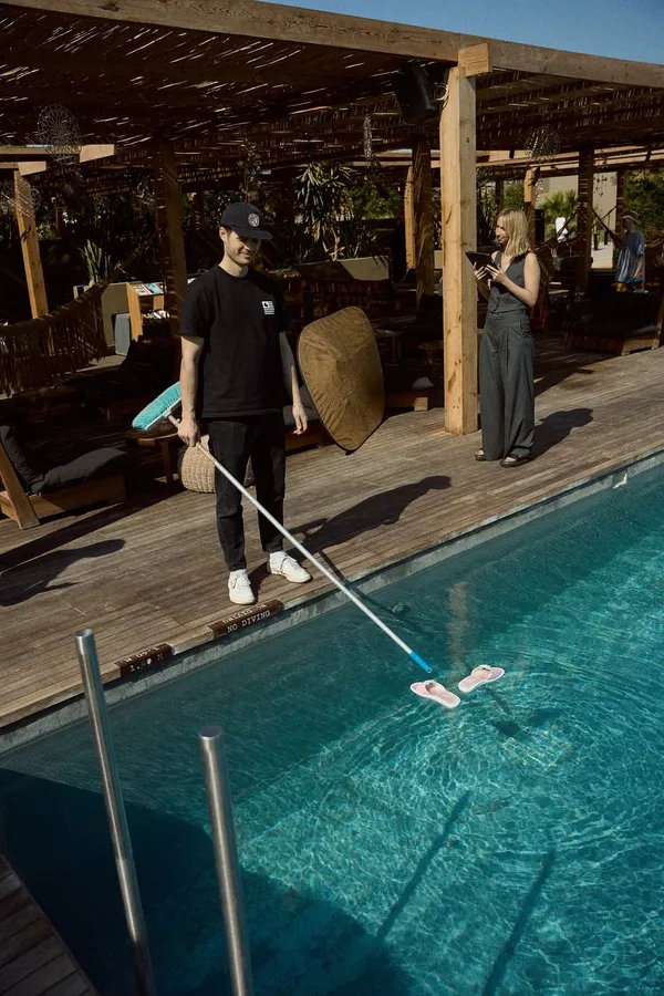 Man cleans a pool beneath a wooden pergola, a woman with a phone nearby, flash by Kilian Amrehn