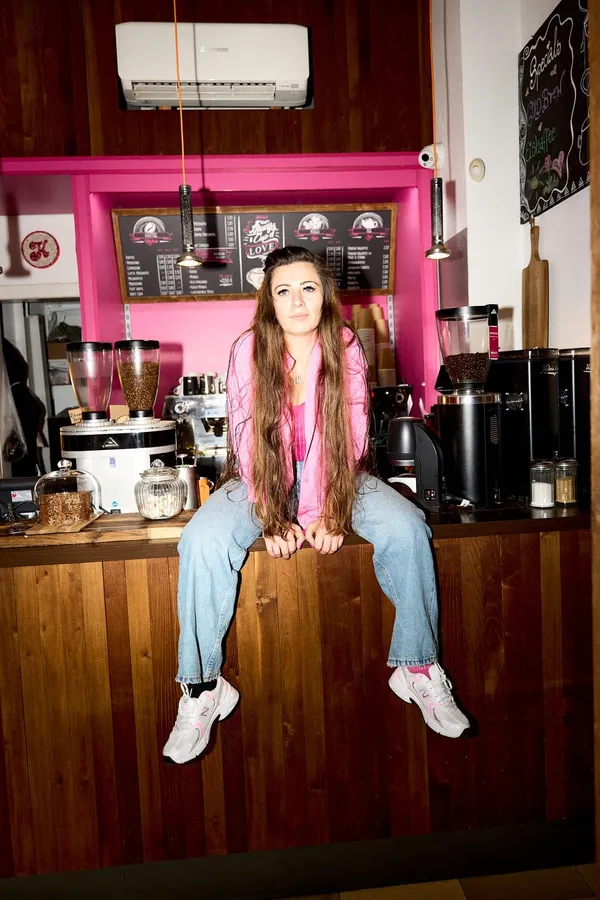 Long-haired woman sits on the wooden counter of a café with a pink wall, flash by Kilian Amrehn