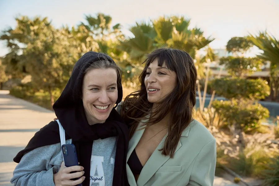 Two laughing women in warm evening light in front of palms and a garden path, flash by Kilian Amrehn