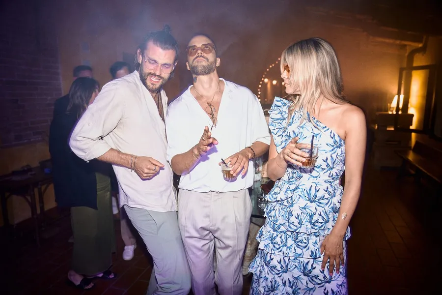 Party guests in light shirts and a floral dress dance in haze and glowing light, flash by Kilian Amrehn